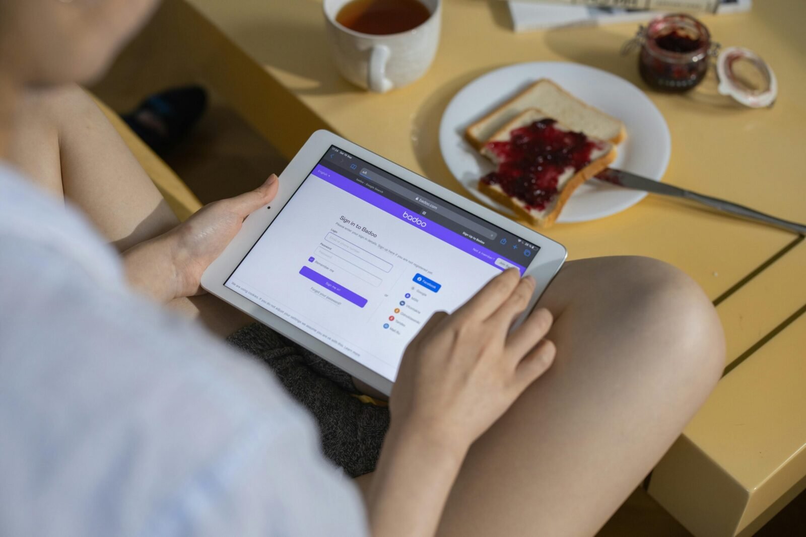 A woman using a tablet while having a snack with jam on toast and tea, reflecting a cozy indoor setting.